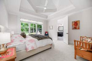 A bedroom with a light color scheme features a bed with floral bedding, a wooden bench, and a furry throw. There's a large window with views of a green landscape. The ceiling has tray design and a fan, and an open door leads to a bathroom.