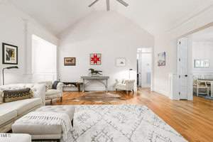 Spacious living room with vaulted ceiling, hardwood floors, and a large area rug. There are two armchairs and a sectional sofa. Wall art and a console table with a decorative horse statue are visible. An open doorway leads to another room.