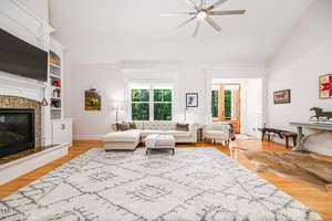 A spacious living room with a large patterned rug on wooden flooring. It features a white sofa, chaise lounge, and ottoman. A modern fireplace and TV are on the left. Large windows let in natural light. Ceiling fan and decorative items complete the space.