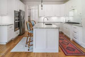 A modern kitchen featuring white cabinetry and a marble island with a built-in sink. Two wicker chairs are placed by the island. There are stainless steel appliances, a tiled backsplash, and a red patterned rug on the wooden floor.