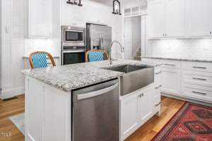 Modern kitchen with a large island featuring a granite countertop and an undermount sink. Stainless steel dishwasher and double oven are visible. White cabinets line the walls. Two blue and white chairs are placed at the island.