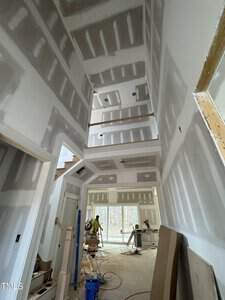 Interior view of a partially constructed house with drywall installed. A tall, narrow hallway with visible beams and an unfinished staircase. Construction tools and materials are scattered on the floor. Natural light enters through large windows.