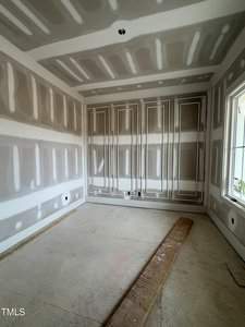 A room under construction with unfinished drywall on the walls and ceiling. The floor is bare, with a wooden plank resting on it. A large window is on the right side, allowing natural light into the space.