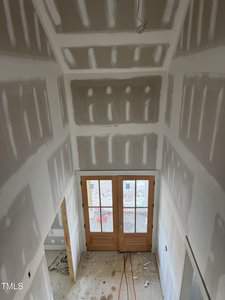 View of an unfinished room with drywall installed and taped, showing double glass doors leading outside. The floor is covered with debris and scattered construction materials, and red extension cords lie on the ground.