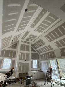 A room under construction with unfinished drywall covering the walls and ceiling. Two windows provide natural light. There's a saw on a stand and various construction materials scattered around, including a ladder and boxes.