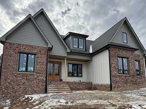 Located at 1401 Cottondale Lane in Wake Forest, this two-story brick and wood house is under construction. It features multiple gabled roofs, large black-framed windows, and a wooden front door. Snow blankets the ground around the unfinished stairs beneath a cloudy sky.