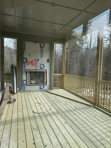A wooden deck with a roof and railing is under construction. There's a covered fireplace with insulation material visible on one wall. The deck overlooks a wooded area, and construction materials are scattered on the floor.