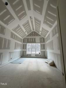 A room under construction with drywall installed on walls and ceiling. The space features a high ceiling and a window at the far end. Two pillows rest on the floor, and there is a small rug to the left.