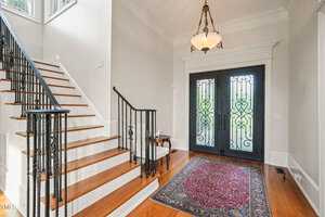 A foyer with a grand iron and glass double door, a staircase with wooden steps and black metal railing on the left. A red patterned rug lies on the wooden floor. An elegant chandelier hangs from the ceiling, and light streams through large windows.