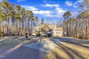 A two-story house with a sloped roof sits in a wooded area. It has beige siding, a three-car garage, and a driveway leading to it. The front yard is grassy with some shrubbery, and tall trees are visible in the background under a blue sky.
