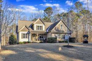 A suburban house with a large front lawn. The house has a beige exterior with stone accents, a dark gray roof, and a small porch. A leafless tree and a decorative wooden structure are in the foreground. Trees are in the background under a blue sky.