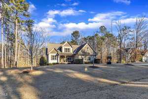 A two-story house with a gray roof and beige exterior sits in a landscaped yard with sparse trees. A paved driveway leads to the front entrance. The sky is partly cloudy, and the surrounding area is wooded.