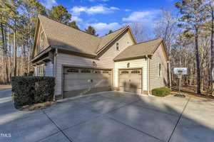 A two-story house with a beige exterior and a three-car garage is surrounded by trees. The driveway is concrete, and a basketball hoop is installed near the garage. The sky is mostly clear with a few clouds.