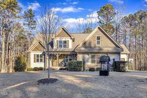 The two-story house at 3667 Jordan Circle in Franklinton boasts tan siding and a dark brown roof, centered on a neatly maintained lawn. In the front yard, a leafless tree and small well feature adorn the picturesque scene, set against tall trees under a clear blue sky.
