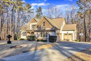 A two-story house with a stone and wood facade is situated in a wooded area. The front yard has minimal landscaping, with a few shrubs and a manicured lawn. The house features a double garage and a paved driveway leading to the entrance.
