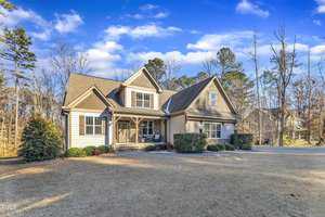 A two-story house with a gable roof and beige siding stands surrounded by a lawn and bordered by shrubs. The sky is partly cloudy, and leafless trees are in the background.