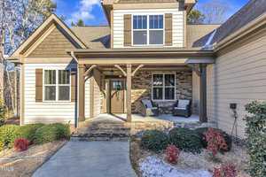 A modern house with a gabled roof and a mix of siding and stone facade. The entrance features a wooden door with glass panels and a small porch with two chairs. Surrounding the house are small shrubs and a stone pathway.