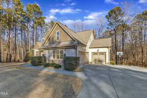 A suburban house with a gabled roof and attached garage. The exterior is light-colored with darker accents. The front yard has trimmed bushes and a wide driveway. Tall trees surround the property under a clear blue sky.