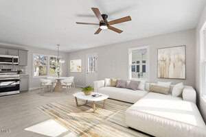 A modern living room at 124 W College Street in Oxford features a large white sectional sofa, a rectangular coffee table on a striped rug, and a ceiling fan. In the background, the dining area boasts a round table with four chairs. Light-colored walls and numerous windows brighten the space.