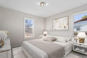 A bright bedroom with a large bed featuring beige and white bedding. Two windows allow natural light. A modern light fixture is on the ceiling, and a neutral-toned abstract painting hangs above the bed. A dresser and two bedside tables complete the room.