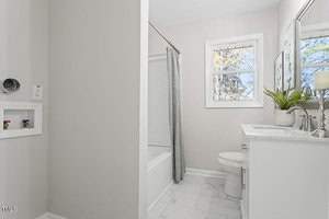 A modern bathroom with light gray walls features a white bathtub with a gray shower curtain, a white vanity with a sink, and a mirror above. A small window lets in natural light. The flooring is white tile. A potted plant and candle sit on the counter.