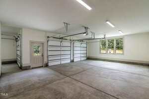 A clean, empty two-car garage with two closed overhead doors, a side entry door, concrete floor, white walls, ceiling lights, and a window letting in natural light.