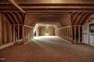 Unfinished attic space with exposed wooden framing and beams, plywood flooring, and a single window at the far end letting in natural light. Some insulation materials are stacked on the left.