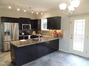 Modern kitchen with dark cabinets, stainless steel appliances, and a granite countertop island. Track lighting and a hanging light fixture provide illumination. A glass door on the right leads to an outdoor area, and a window is above the sink.