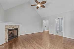 A spacious room with polished hardwood floors, a large stone-framed fireplace, and a vaulted ceiling. A ceiling fan with a light hangs above. There are two doorways leading to other rooms with light-colored walls.
