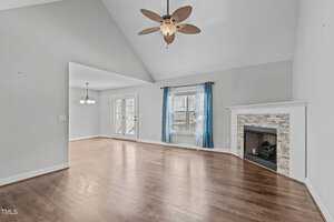 A spacious room with light gray walls and a wooden floor. It features a ceiling fan with a light in the center, an empty fireplace with a tiled surround, and windows with sheer blue curtains. Double glass doors open to an outdoor deck area.