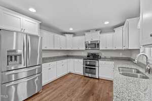 A modern kitchen with stainless steel appliances, including a refrigerator, stove, and microwave. White cabinetry lines the walls, and a granite countertop includes a sink with a curved faucet. The floor is made of polished wood.