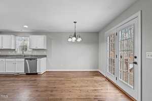 Bright kitchen and dining area with hardwood floors. White cabinets and stainless steel dishwasher are on the left. A hanging light fixture is centered above the dining space. Double glass doors lead to an outdoor area on the right.