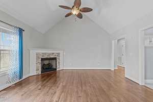 A spacious, unfurnished room with wooden floors and light-colored walls. The room features a fireplace with a stone surround, a ceiling fan with wooden blades, and a window with a blue curtain. The ceiling is vaulted.