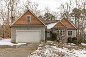 This charming single-story house at 4294 Sustain Circle, Franklinton, boasts gray siding with brick accents. It features a two-car garage and a small porch. The roof, covered in shingles, is framed by bare trees. A light snow dusts the driveway, flanked by shrubs near the entrance.
