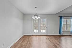 A room with wooden flooring and a hanging chandelier. There is a glass door leading outside and a window with blue curtains. Walls are painted light gray, and the room appears empty. Natural light is coming in through the glass door and window.