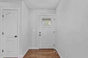 A small hallway featuring a light gray front door with a windowpane and a white interior door on the left. The floor is made of wooden planks, and there is a light switch on the wall.