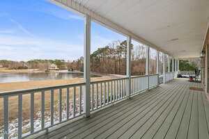 A long wooden porch with white railings overlooks a serene body of water, surrounded by trees and a grassy area. A snow-dusted landscape is visible under a clear blue sky. A few chairs are placed along the porch.