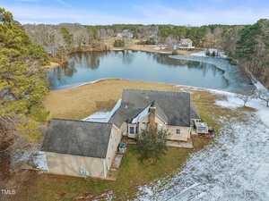 Aerial view of a beige house with a dark roof, situated by a large pond surrounded by grass and trees. Snow covers parts of the ground. The landscape includes a cluster of trees and several other houses in the distance.