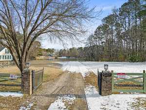 A gravel driveway leads through a gate with brick pillars on a snowy day. There are fields on either side, and a pond is visible in the distance. A house is partially seen to the left, and trees line the right side of the driveway.