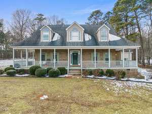A two-story brick house with a wraparound porch, featuring multiple gabled dormer windows and a green front door. The yard has some snow patches, and tall trees are in the background.