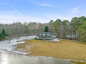 A large house at 4056 Fairfield Court sits on a grassy field surrounded by trees. A light dusting of snow covers parts of the ground and the roof. A small body of water is visible in the foreground under the clear, blue Youngsville sky.