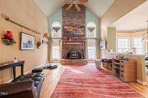 A spacious living room with high ceilings features a brick fireplace flanked by tall windows. A red-striped rug covers the wooden floor. Various decorative items are displayed on shelves, and a ceiling fan is centered above the room.