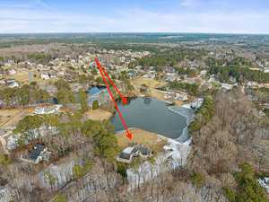 Aerial view of a residential area with scattered houses and a large pond surrounded by trees. Two red arrows point from a house in the foreground to another house across the pond. The landscape features mixed woodland and open fields.