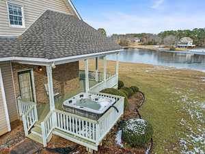 A wooden deck attached to a house features a hot tub under a covered roof. The house has a grassy yard with patches of snow and borders a lake with trees and houses across the water.