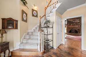 A staircase with white carpet ascends in a residential hallway. The walls are light yellow with two floral paintings. Shelves with plants are near the stairway. A doorway leads to a room with a fireplace and hardwood floor.
