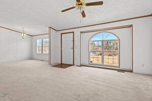 Spacious living room with white walls and beige carpet. Features a central ceiling fan, an arched window on the right, and a doorway with sidelights. A small dining area with a hanging light is visible in the background.