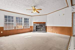 A room with white walls and a ceiling fan. Three large windows are on the left wall, and a stone fireplace with a black hearth is centered on the far wall. The floor is carpeted, and there is wood paneling along the lower portion of the walls.