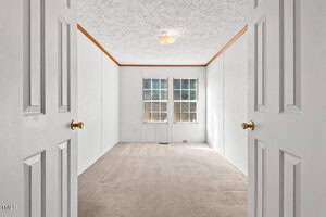 A small, empty room with white walls and beige carpet. The ceiling has a textured finish. Two closed white double doors are at the entrance. There are two large windows on the far wall, allowing natural light into the space.