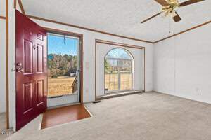 A room with a red door, open to the outside. It has a beige carpet and white walls. There is a large arched window with a view of trees. A ceiling fan is mounted above. The exterior landscape includes grass and trees.