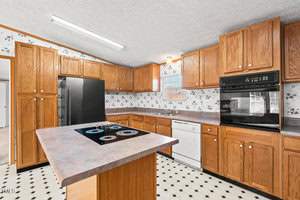 A spacious kitchen with wooden cabinets, a black oven, a dishwasher, and a large refrigerator. An island with an electric stovetop is in the center. The floor has a black and white checkerboard pattern, and the walls feature floral wallpaper.